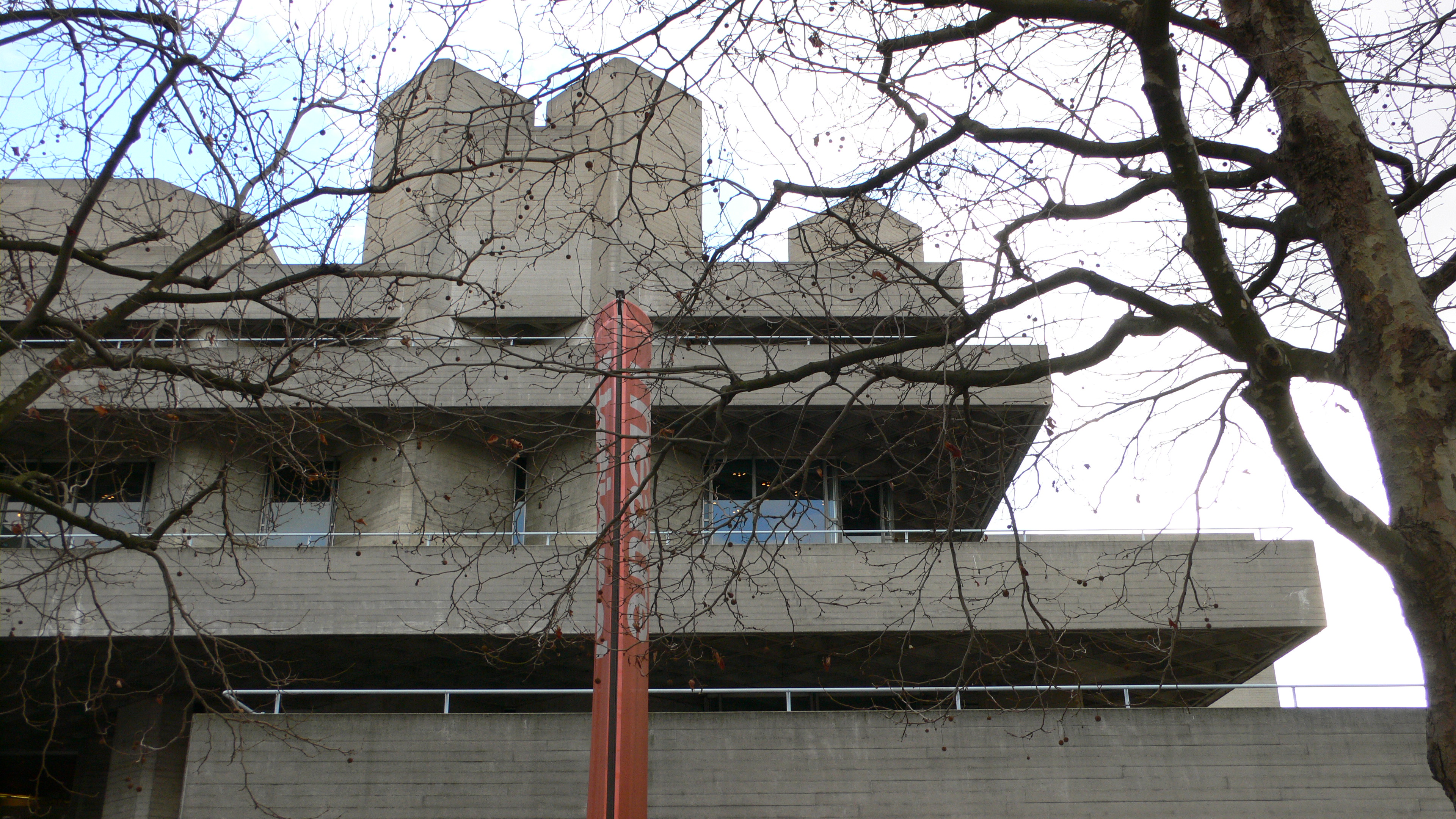 Cantilevered concrete terraces of the National Theatre seen through bare branches.