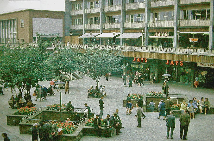 People walk, sit and gather around planted beds in Coventry’s Upper Precinct, beneath two levels of shopfronts, in 1962.