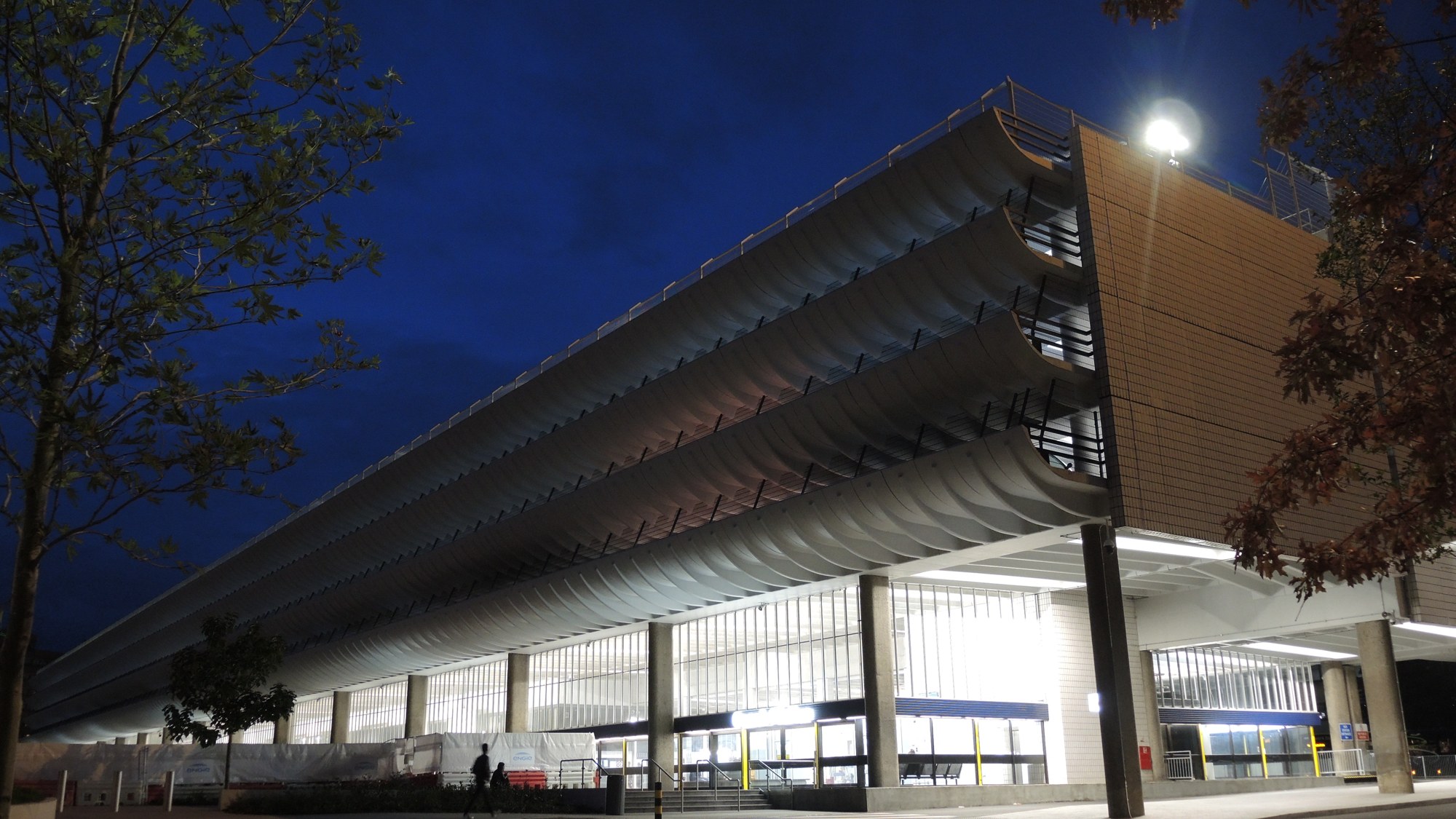 Preston Bus Station at night: curved concrete parking decks rise above a brightly lit ground-floor concourse, with a small figure below.